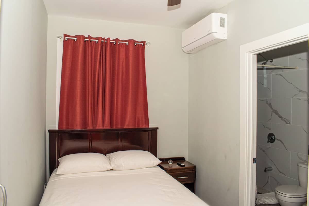A small, neatly arranged bedroom with a single bed, a nightstand, a red curtain, and a bathroom with a toilet and shower.