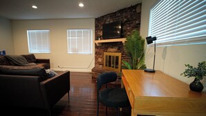 A living room with a sofa, a wooden table, a TV mounted on a brick wall, and two windows with blinds. - Beautiful & Centrally Located close to everything 3- bedroom home, AC, WiFi (Baldwin Park)