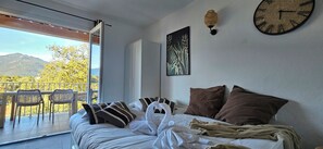 Habitación de hotel con cama, almohadas, manta, un reloj de pared, un cuadro y un balcón con sillas y vistas a las montañas.