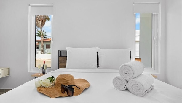 Una habitación de hotel con una cama, ropa de cama blanca, un sombrero de paja, gafas de sol y toallas enrolladas.