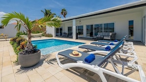 Un área de piscina moderna al aire libre con sillones de descanso, una palmera y un patio con mesa y sillas.