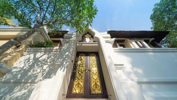A grand entrance with a dark wooden door and intricate golden designs, flanked by white walls and a tree providing shade. - Luxurious Pattaya Pool Villa (Sattahip)