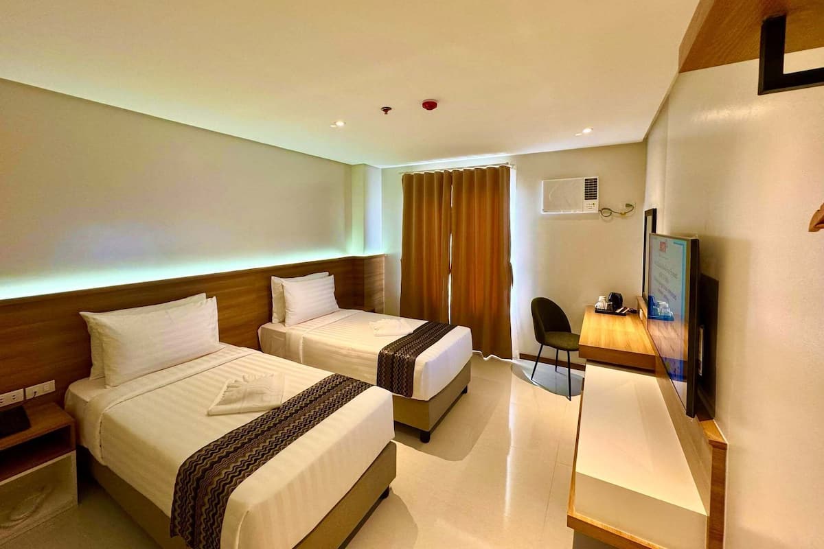 A hotel room with two beds, a desk, a chair, a TV, and an air conditioning unit.