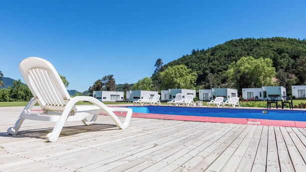 A poolside area with white lounge chairs, a blue pool, and a forested hill in the background.