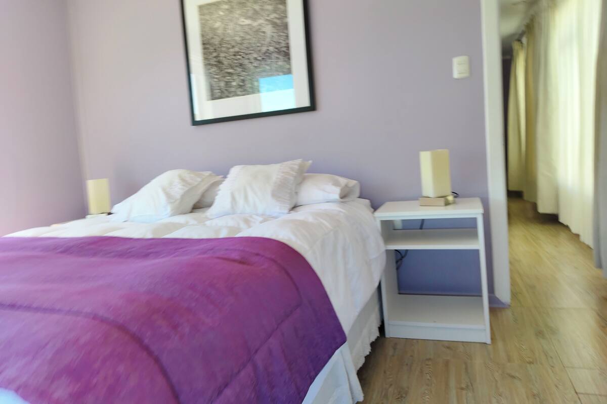 Une chambre à coucher avec un lit, des oreillers blancs, une couverture violette, une table de chevet avec une lampe et un tableau encadré accroché au mur.