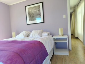Un dormitorio con cama, almohadas blancas, una colcha morada, una mesita de noche con lámpara y un cuadro en la pared.