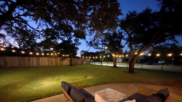 An outdoor seating area with a wicker sofa and cushions, surrounded by a well-lit lawn and trees.