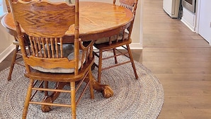 A dining area with a round wooden table and chairs, a kitchen in the background, and a patterned rug on the floor.