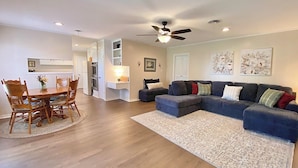 A spacious living room with a large sectional sofa, a dining area with wooden chairs, and a kitchen with white cabinets.