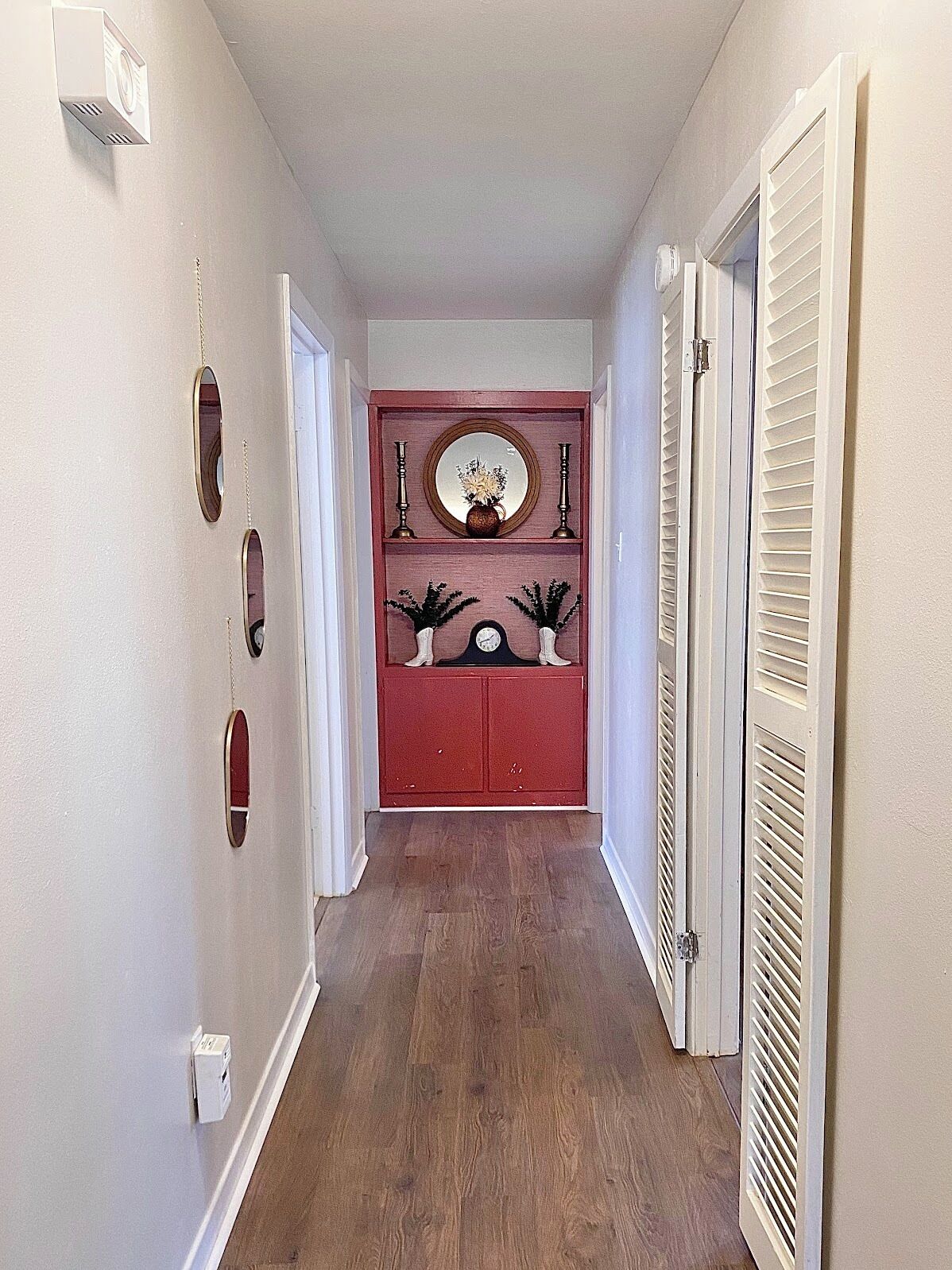 A hallway with a wooden floor, white walls, and a red door at the end. There are three wall-mounted light switches and a white electrical outlet on the left wall.