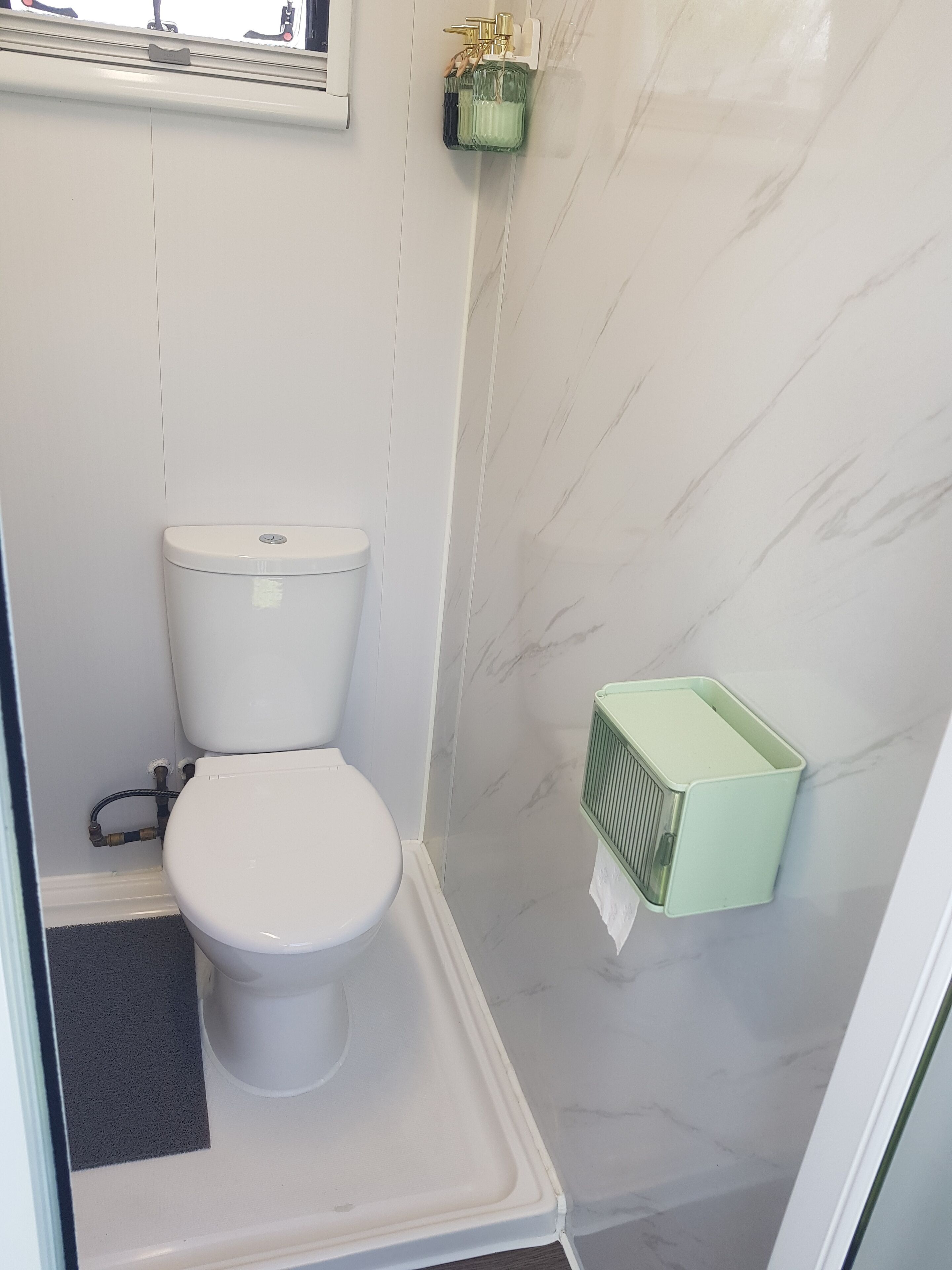 Et lille badeværelse med toilet, en grøn skraldespand og et vindue over toilettet.
