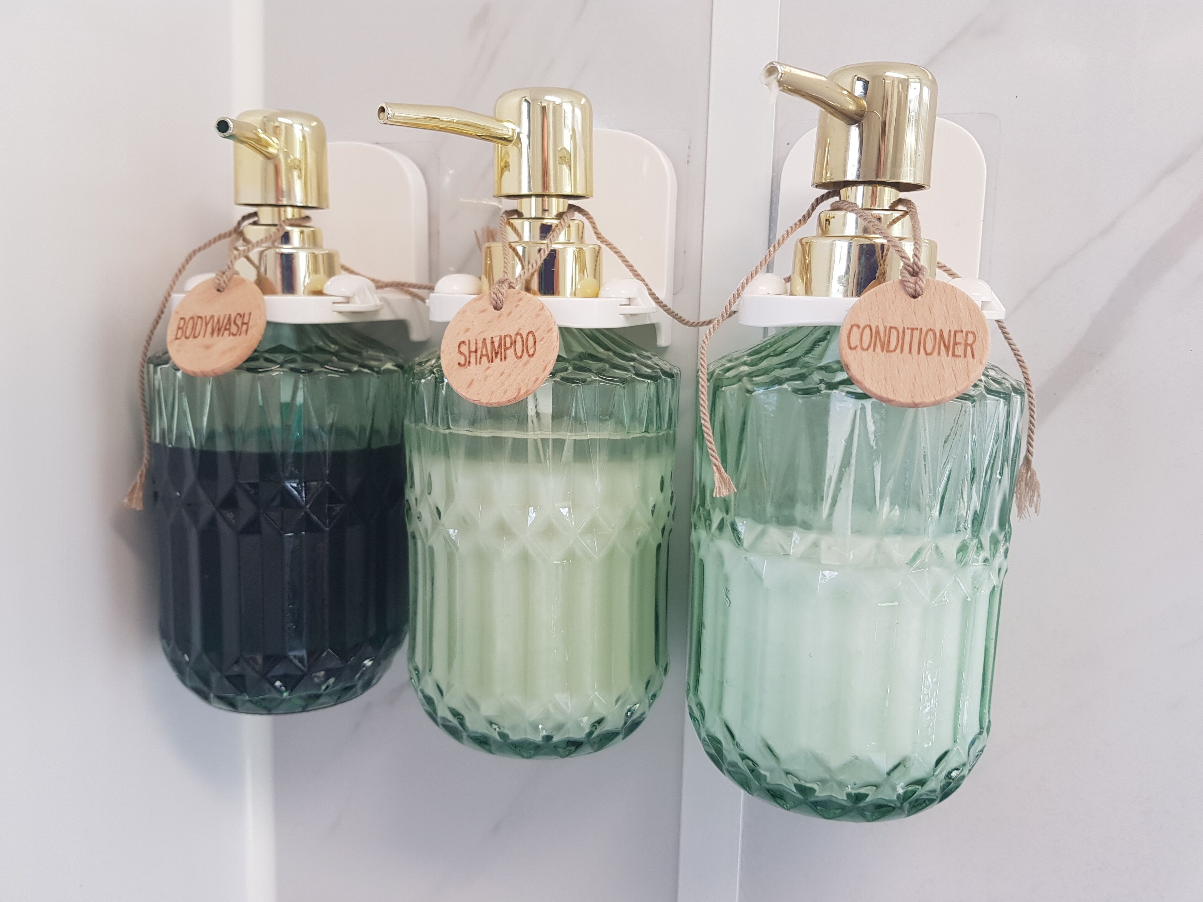 Tre glasflasker med pumper, mærkede 'BODYWASH', 'SHAMPOO' og 'CONDITIONER', hænger på en hvid væg.