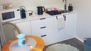 A compact kitchen with white cabinets, a microwave, a kettle, and a sink. There is a small dining table with a blue vase and a white rug. - QUIET COMFY PRIVATE TINY HOME (Caboolture)