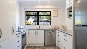 A modern kitchen with white cabinetry, a dishwasher, a stainless steel refrigerator, and a window with a view of a house and greenery. - Bird Songs and lake views with wifi (Acacia Bay, Taupo)