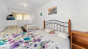 A bedroom with a bed, two white cribs, a wooden nightstand, a window with blinds, and a painting on the wall. - Westhall in Kinloch Taupo (Kinloch)