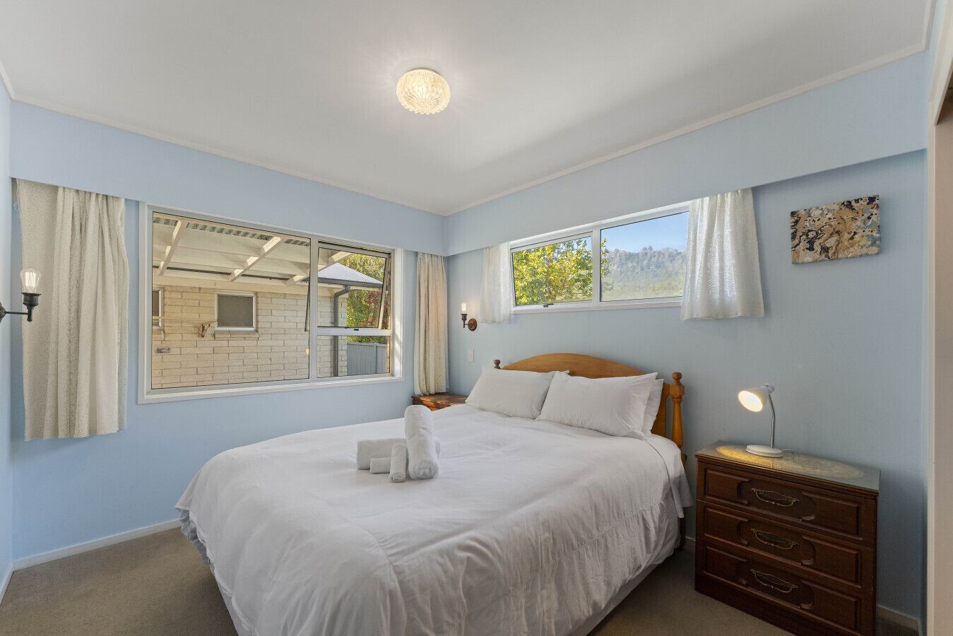 Une chambre à coucher avec un grand lit, deux fenêtres, une table de chevet en bois et une commode.