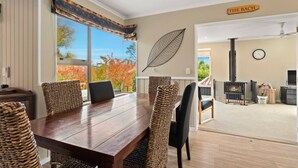A dining area with a wooden table and woven chairs, a large window with a view of autumn trees, a wall-mounted leaf decoration, and a sign reading 'THE BACH'. - Peaceful on Par Kinloch (Kinloch)