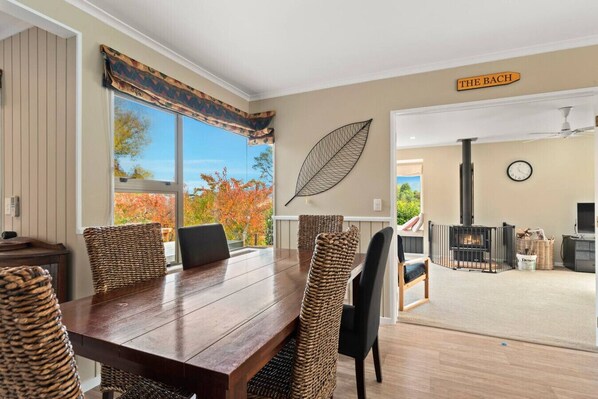 A dining area with a wooden table and woven chairs, a large window with a view of autumn trees, a wall-mounted leaf decoration, and a sign reading 'THE BACH'.