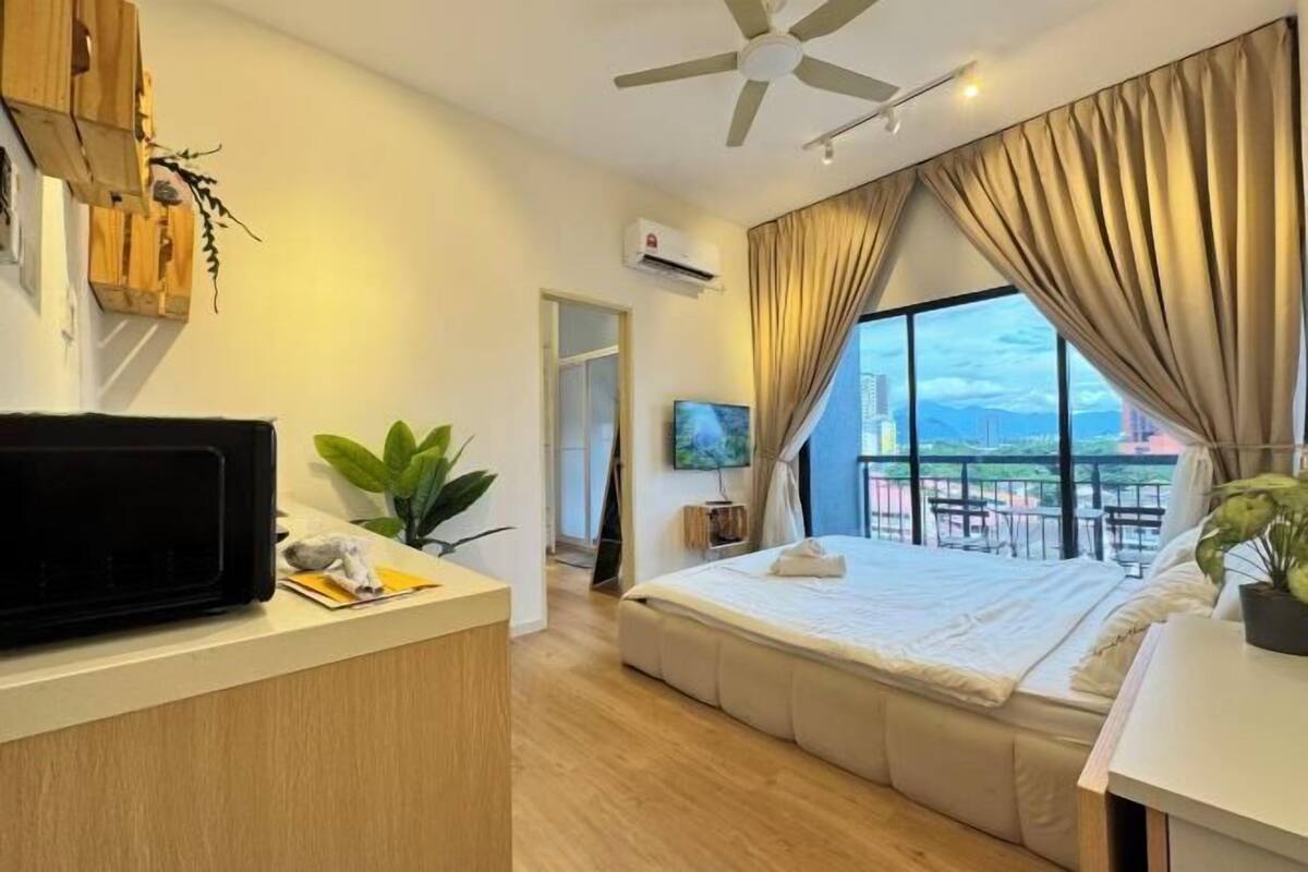 A modern hotel room with a large bed, a desk with a television, a ceiling fan, and a view of a cityscape through large windows.
