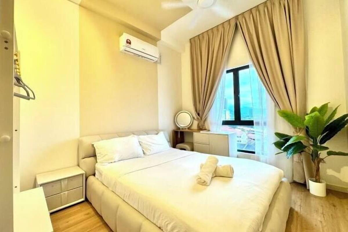 A hotel room with a bed, a bedside table, an air conditioning unit, a window with curtains, and a potted plant.