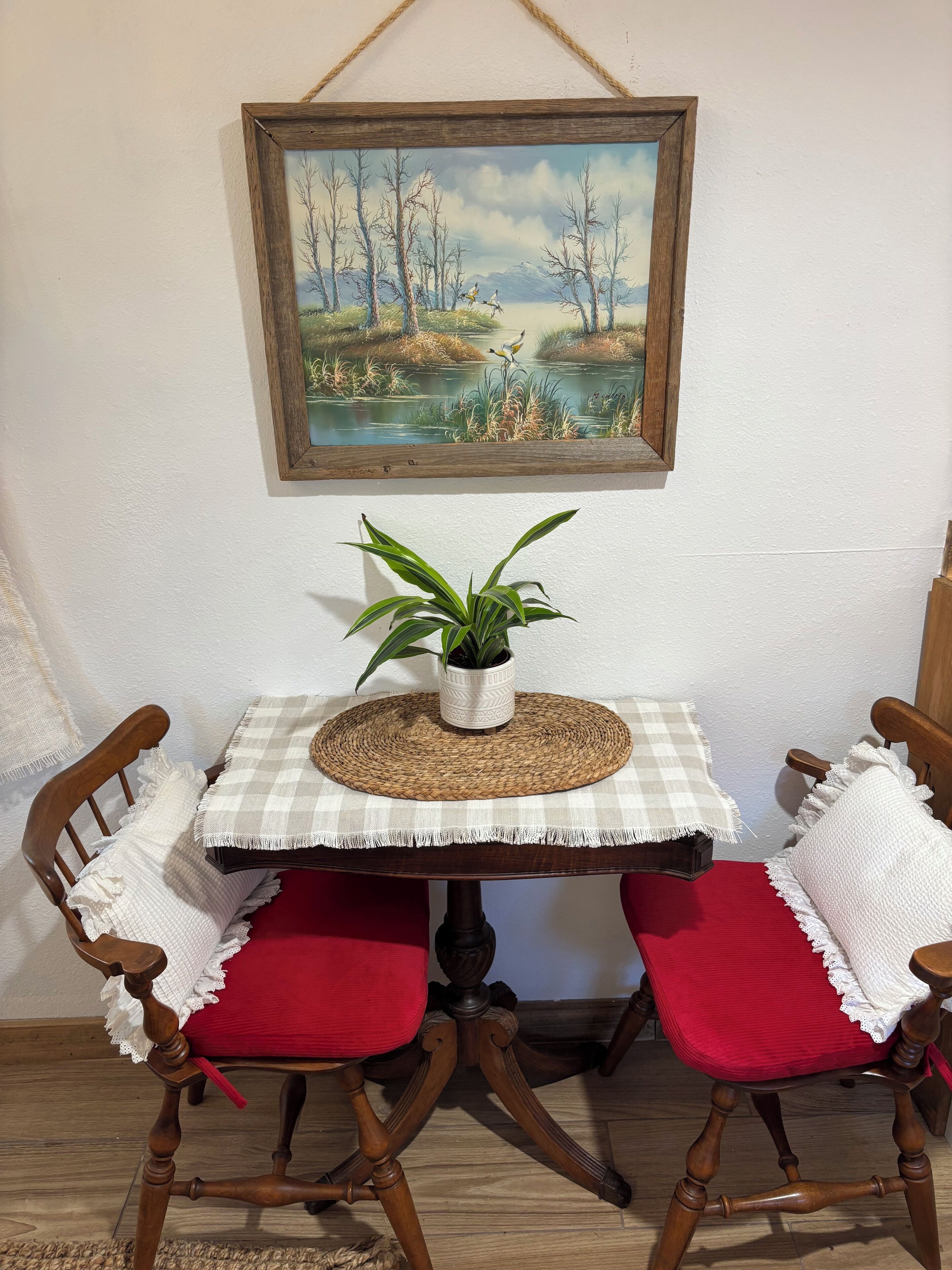 A dining area with a table set for two, featuring a woven placemat, a checkered tablecloth, and two wooden chairs with red cushions. A framed painting of a landscape hangs above the table.