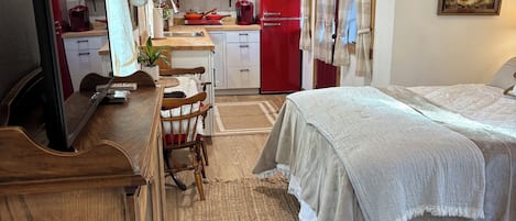 Une chambre à coucher de taille modeste, avec un lit, une commode en bois, un téléviseur et, au fond, un coin cuisine.