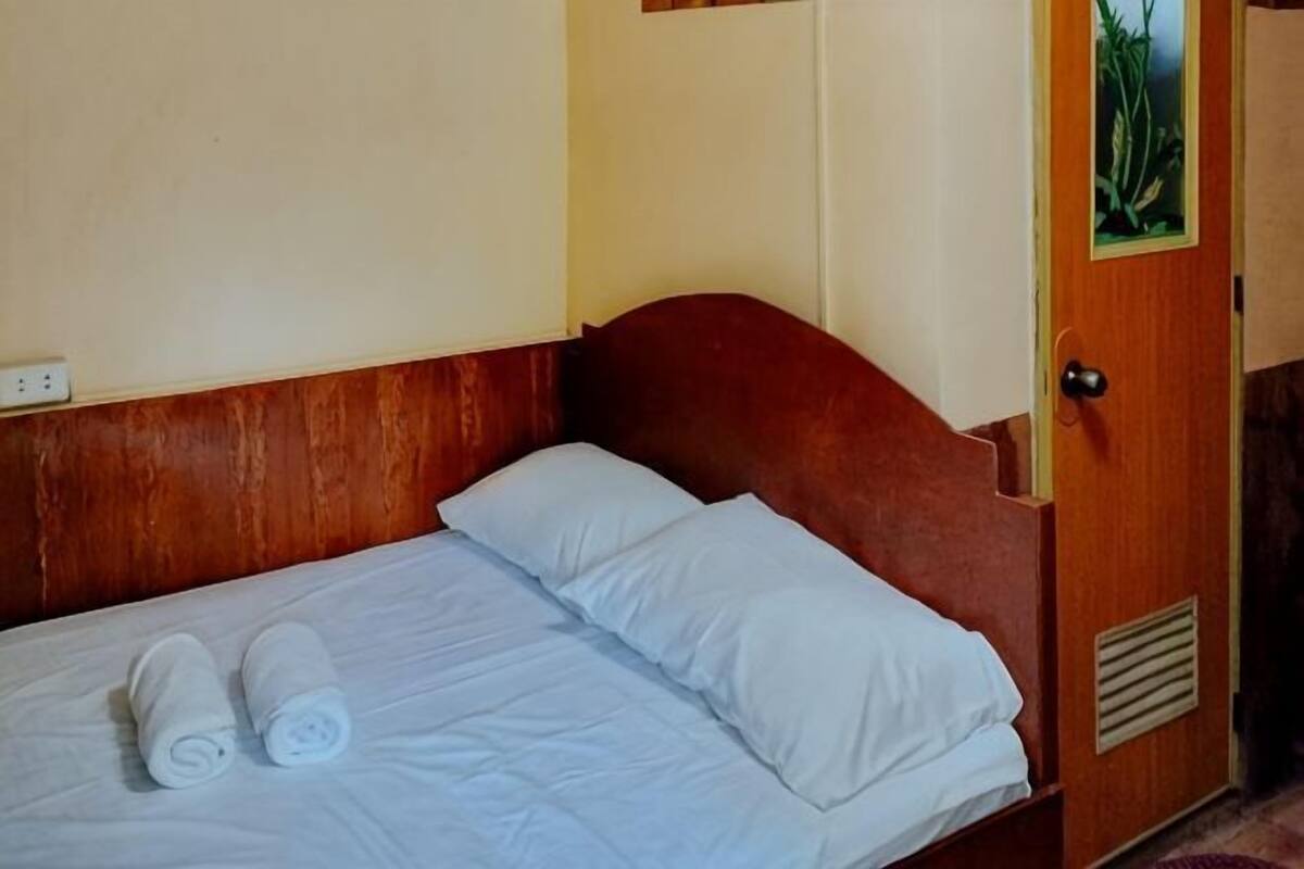 A single bed with white linens and two rolled towels, a wooden headboard, a door with a window, and a power outlet on the wall.