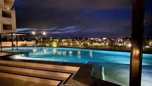 Vista nocturna de una piscina con un bordeado de baldosas y una vista de la ciudad al fondo.