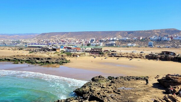 A coastal town with a sandy beach, rocky shoreline, and buildings in the background. - Paradise House Apartment in Tamraght (Tamraght)