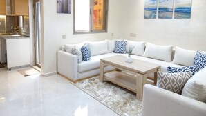 A modern living room with a white sofa set, a wooden coffee table, and a rug. There is a kitchen area with a sink and cabinets visible through an archway. - Paradise House Apartment in Tamraght (Tamraght)