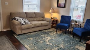 Een woonkamer met een beige bank, een blauwe fauteuil, een blauw vloerkleed en een plafondventilator.