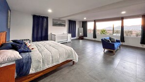 Una habitación de hotel moderna con una cama grande, un área de descanso con un sofá azul, un escritorio blanco y vistas del paisaje a través de ventanas amplias.