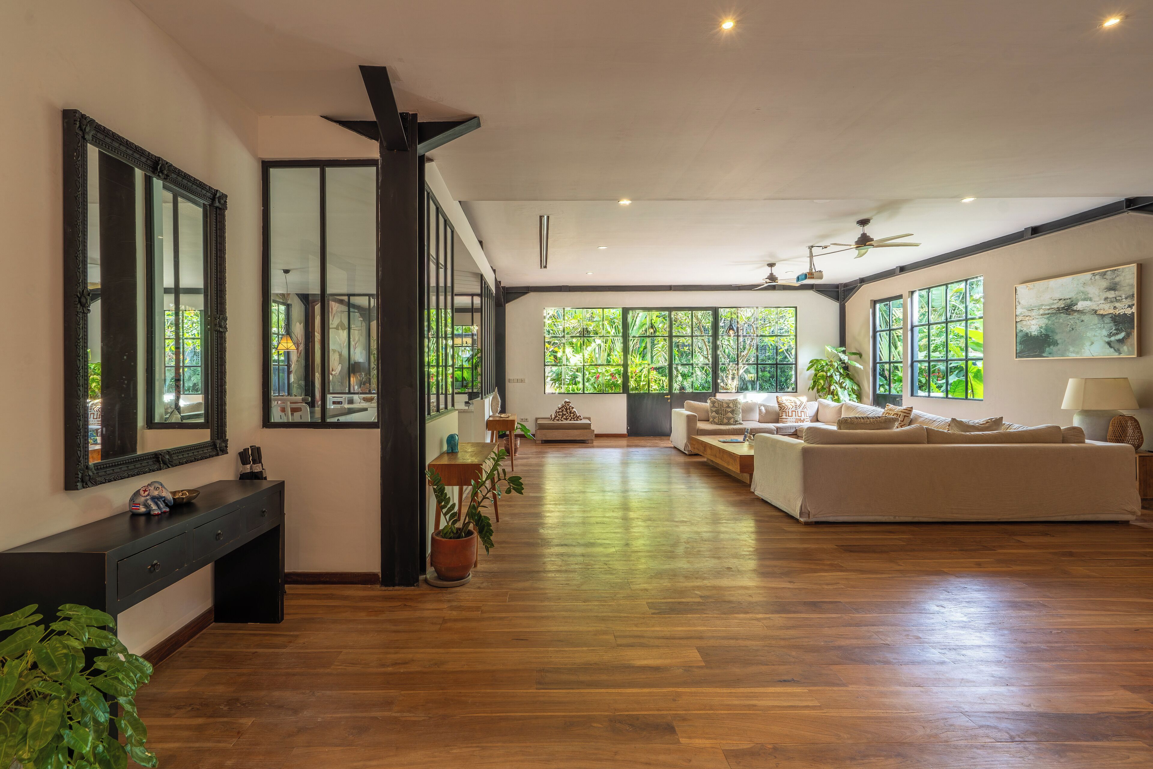 A spacious living area with a large sofa, a coffee table, and a dining table. There are large windows providing natural light and a view of greenery outside.