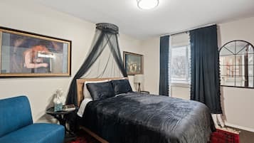 Una camera da letto con un letto, una poltrona blu, un comodino, una lampada, una finestra con tende e un quadro appeso al muro.