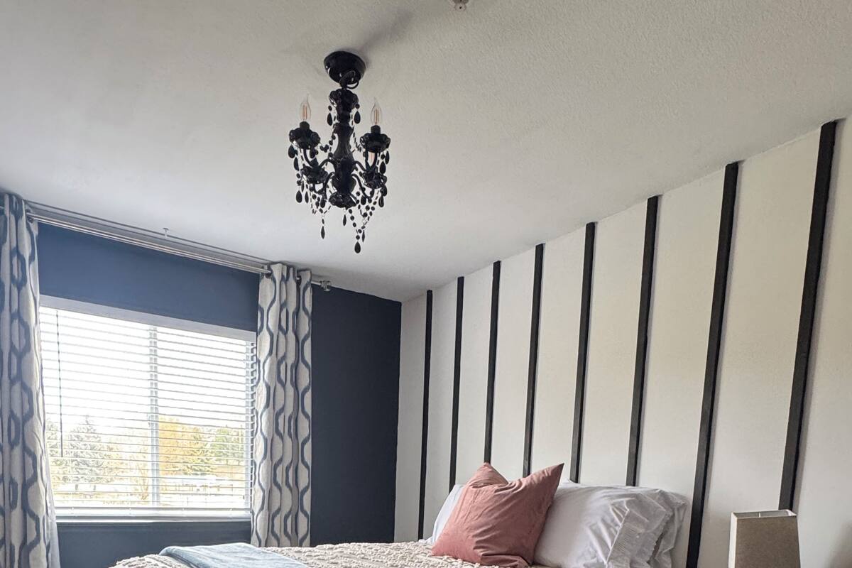 A bedroom with a bed, a nightstand, a lamp, a chandelier, and a window with curtains.