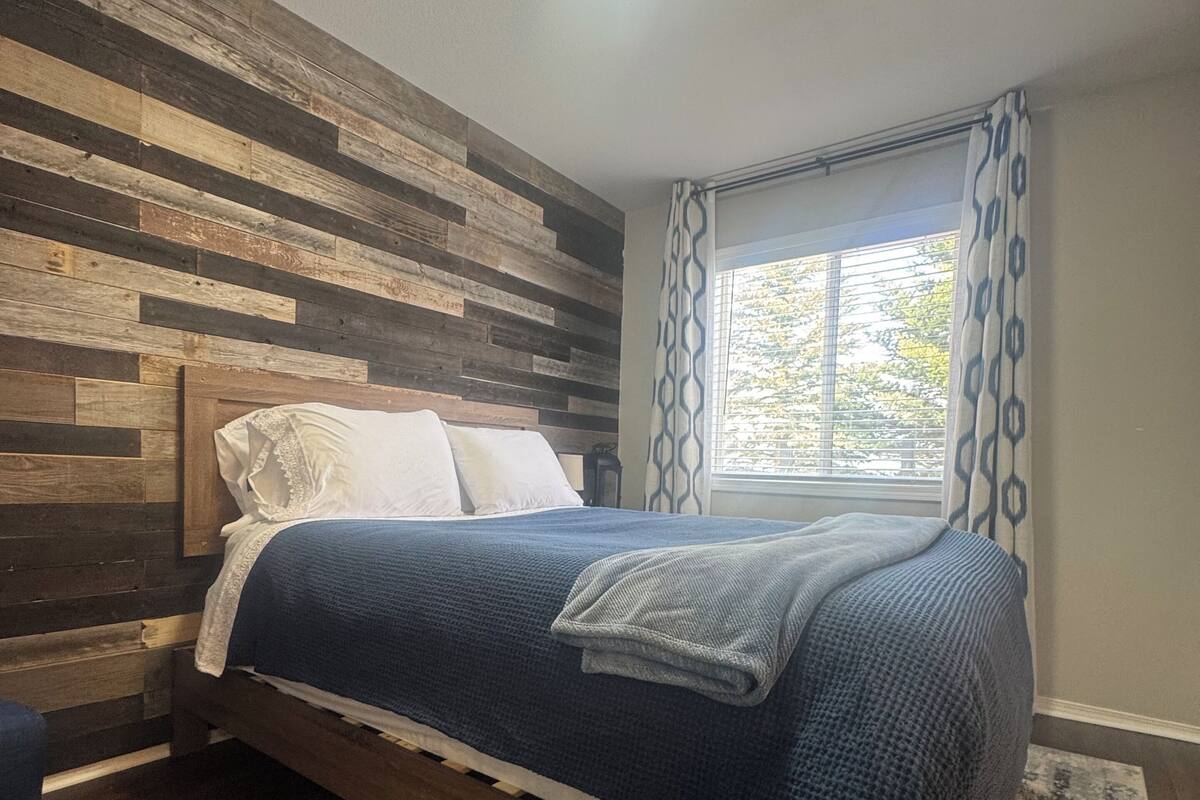 A bedroom with a wooden headboard, a bed with a blue blanket, a window with patterned curtains, and a patterned rug.