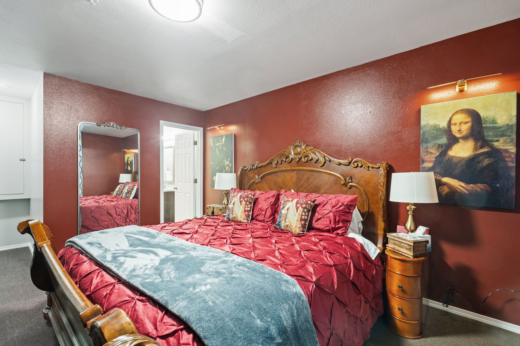 Een slaapkamer met een houten bed, beddengoed in rood en blauw, een spiegel, een kaptafel en een schilderij van de Mona Lisa.