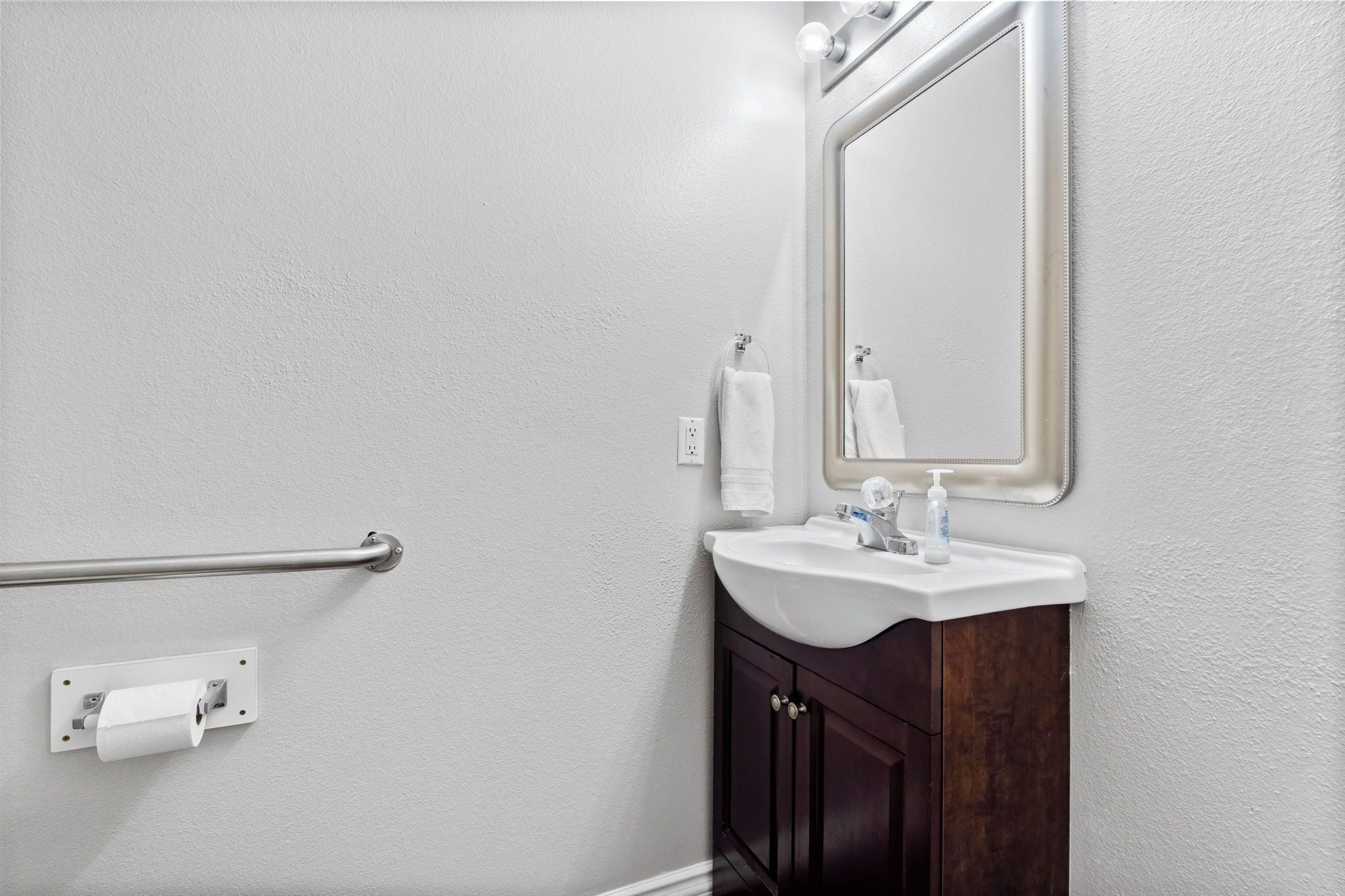 Une salle de bain avec un lavabo blanc, un meuble-lavabo en bois foncé, un miroir et une barre d’appui.