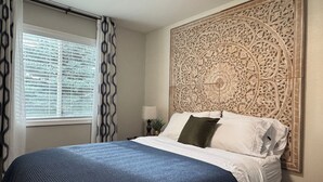 A bedroom with a large bed, a decorative headboard, a window with blinds, and a patterned curtain. - Library INN LLC (Lava Hot Springs)