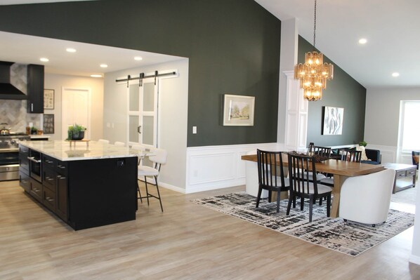 A modern kitchen with a large island, dark walls, and a dining area with a chandelier. - 7BR Retreat - Hot Tub • Fire Pit • 2 King Suites • Near SLC (Sandy)