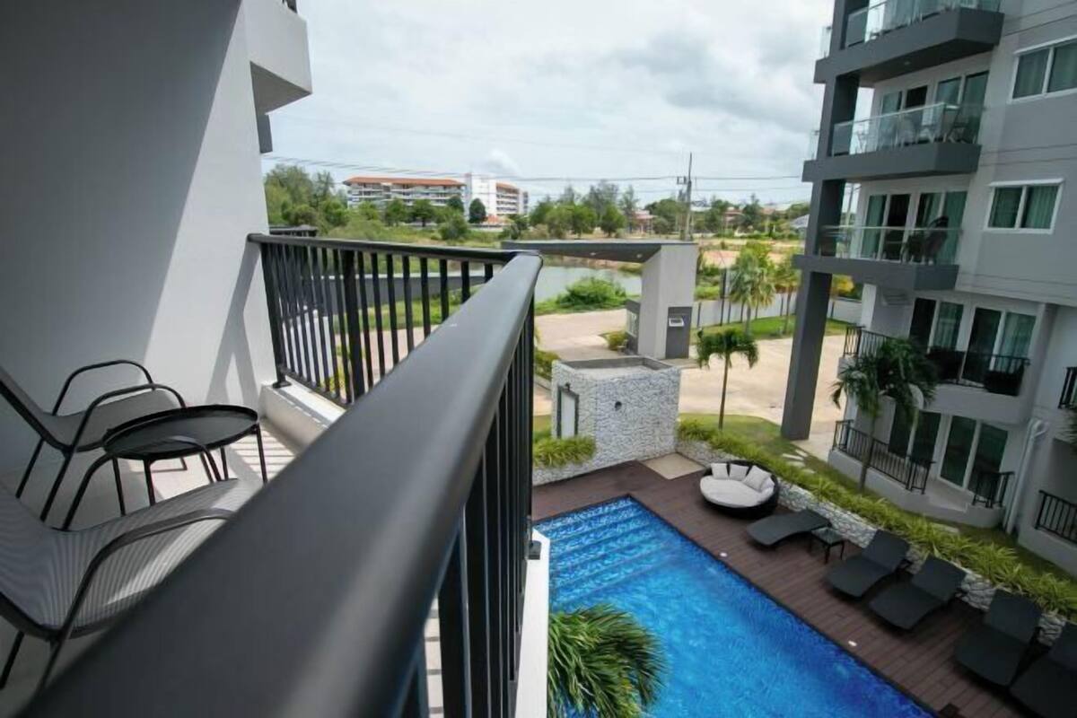 A balcony with a pool, lounge chairs, and a view of a building and greenery.