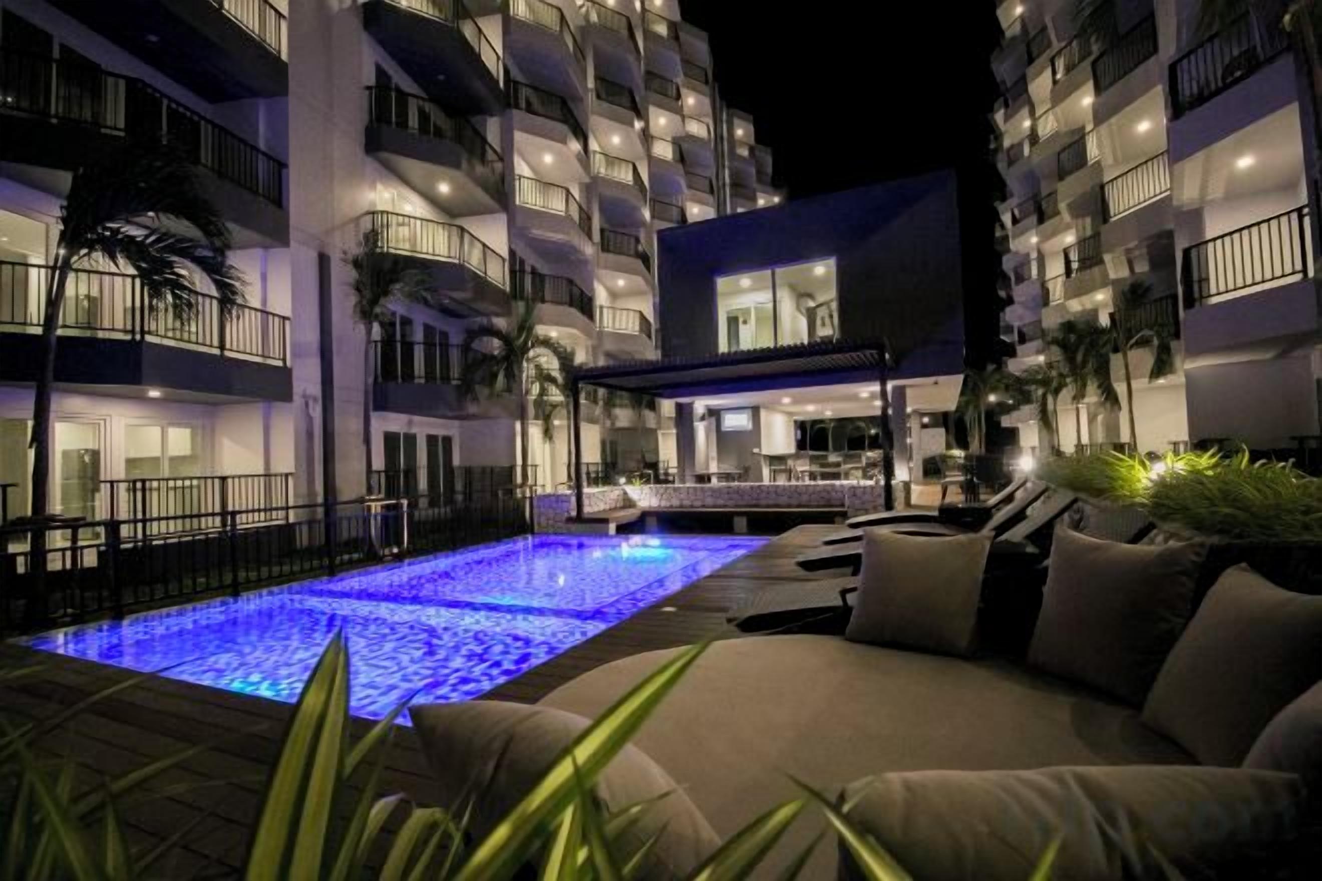 Un espace piscine aménagé avec un canapé et des chaises longues, entouré d’un immeuble à plusieurs étages doté de balcons.