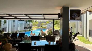 Un área de estar al aire libre techada con vista a la piscina, una pantalla digital y una planta en maceta.