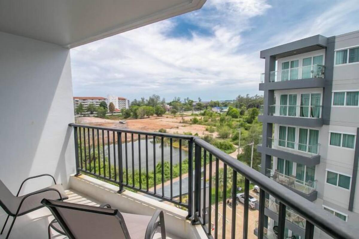 A balcony with a view of a river and surrounding greenery, a modern building with multiple floors, and a clear sky.