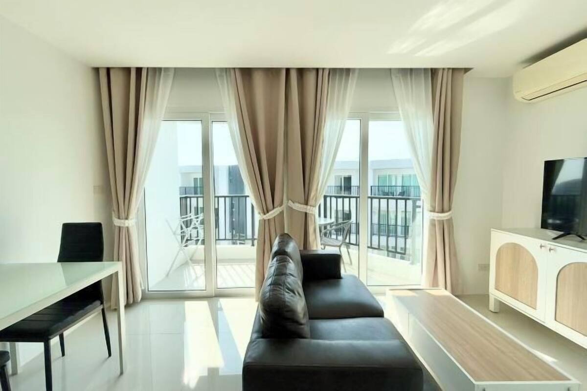 A modern living room with a sofa, TV, and dining area, featuring large windows and a balcony.
