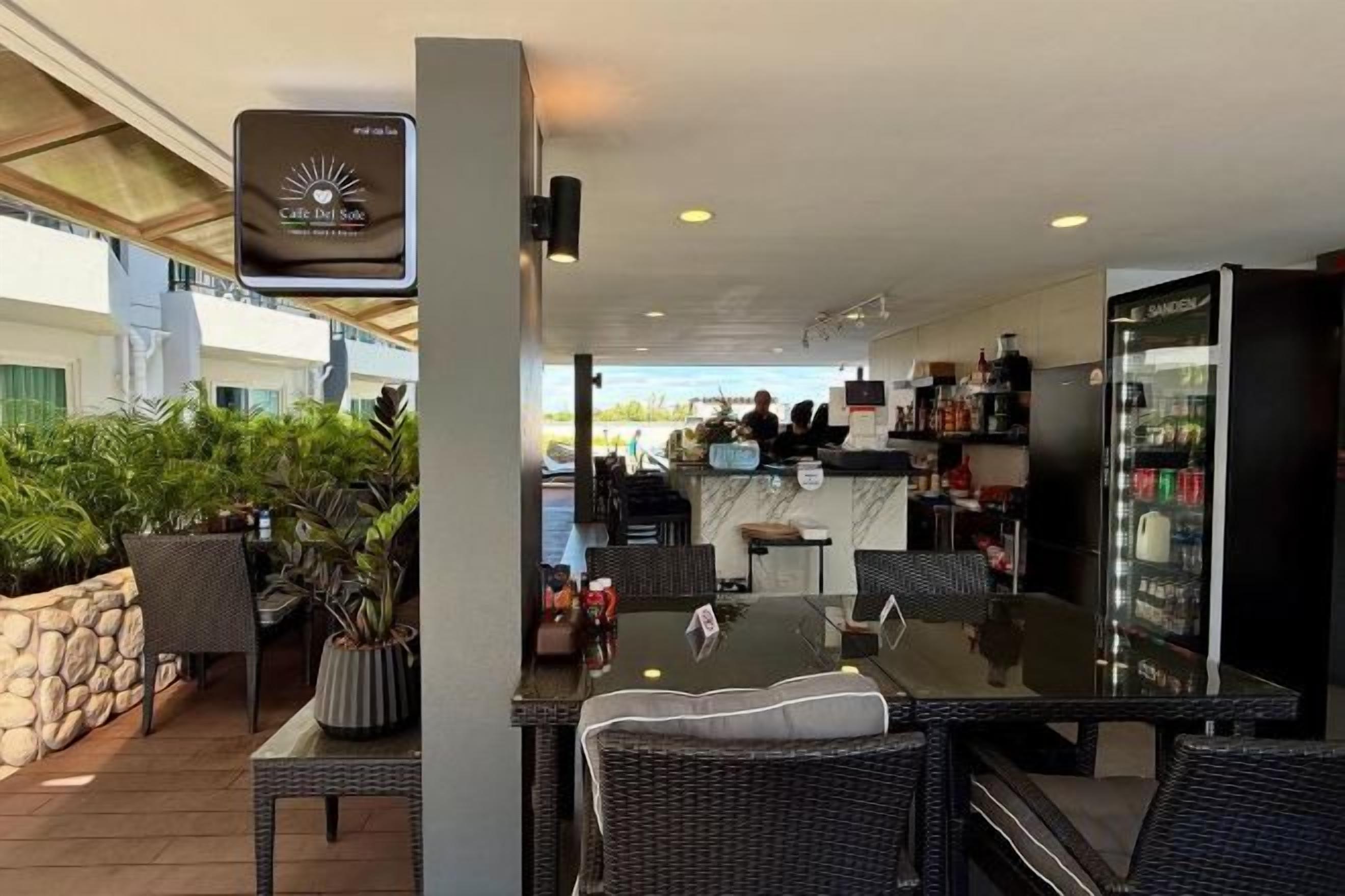 Un área de comedor al aire libre con mobiliario de mimbre, una zona de bar con refrigerador lleno de bebidas y vista a la playa.