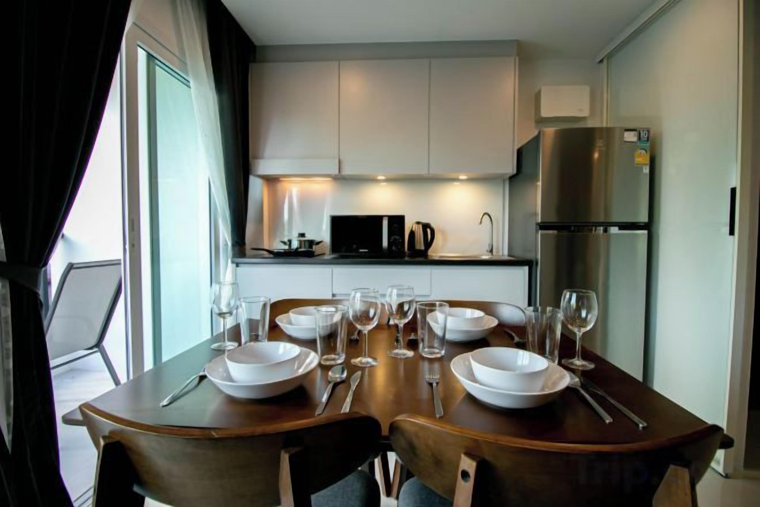 Comedor con mesa lista para cuatro, cocina al fondo y vista por la ventana.