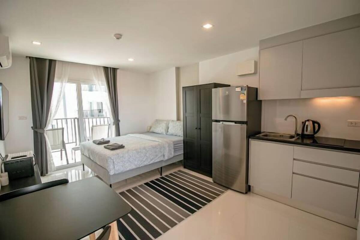 A modern hotel room with a bed, a kitchenette, a refrigerator, and a window with curtains.