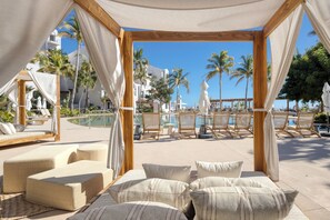 Un área de descanso junto a la playa con asientos acolchados y vistas a la piscina y las palmeras.