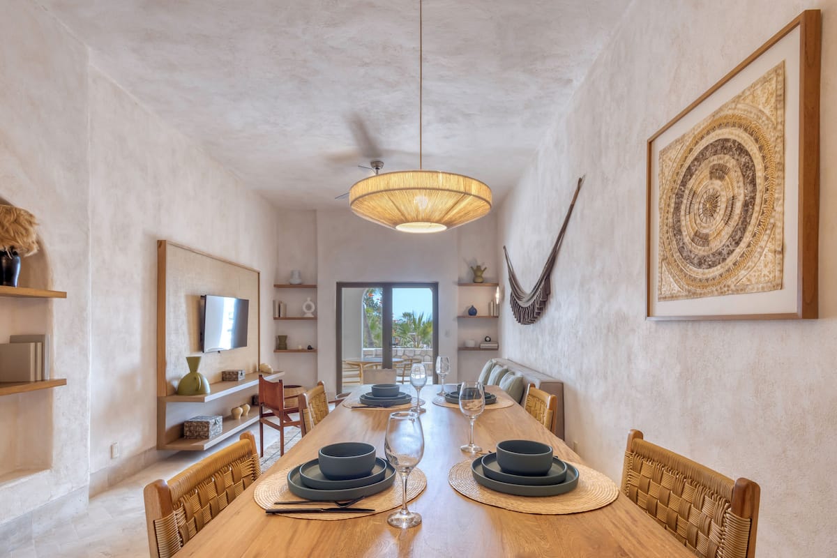 Un comedor con una larga mesa de madera, sillas, una lámpara colgante y una obra enmarcada en la pared.